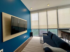 Ein modernes Wohnzimmer mit einem großen Flachbildfernseher an einer blauen Wand, einem dunklen Sofa, einem kleinen Couchtisch und Blick auf eine Stadtlandschaft durch große Fenster.