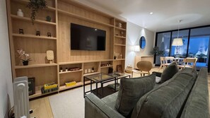Ein modernes Wohnzimmer mit einem großen Flachbildfernseher, eingebauten Holzbögen, einem grauen Sofa und einem Essbereich mit Tageslicht.