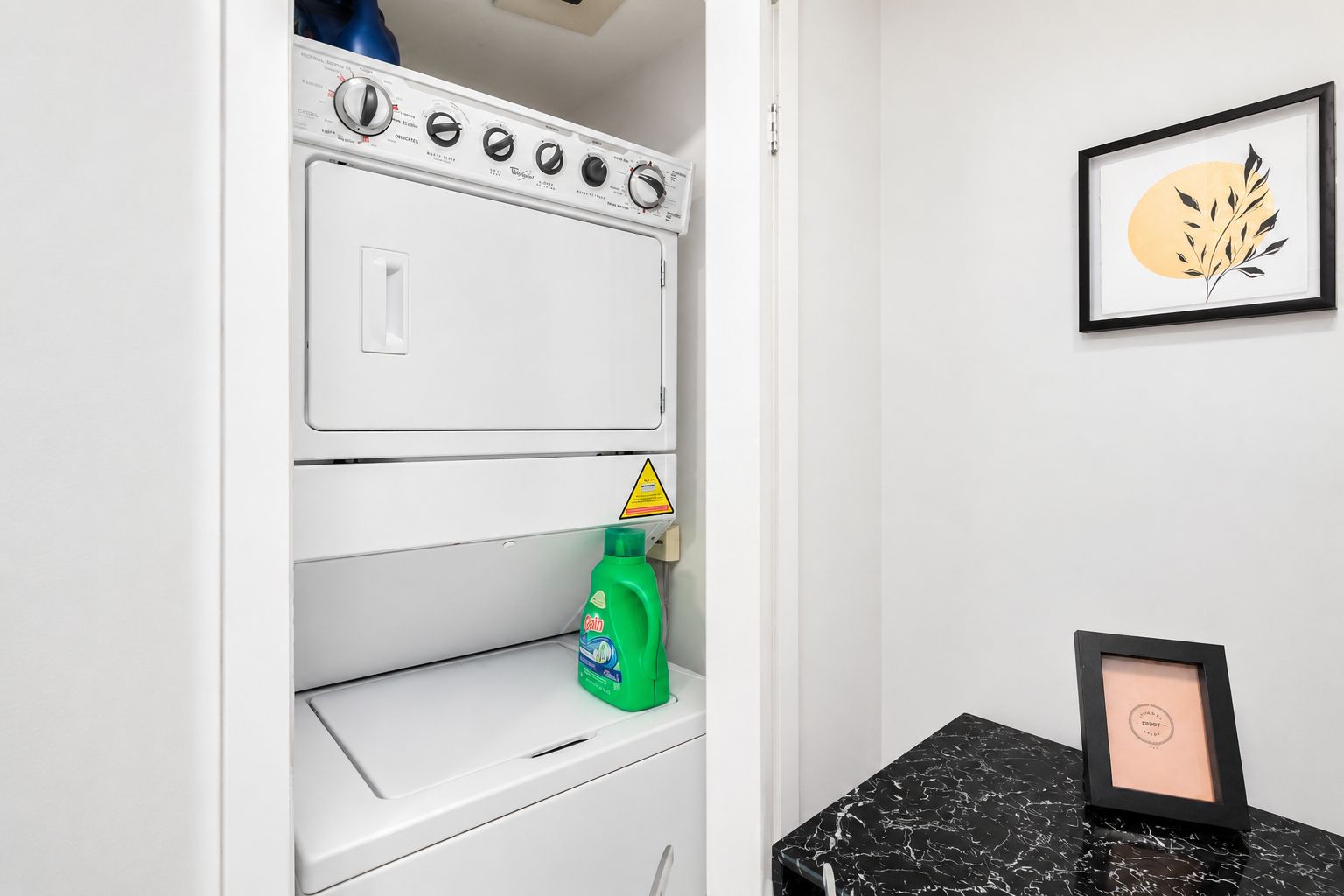 Uma lavanderia com máquina de lavar, um frasco de detergente e um quadro em cima do balcão.