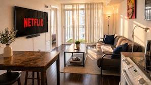 Una sala de estar con un televisor mostrando 'NETFLIX', un sofá, una mesa de centro, una lámpara y una zona de cocina con estufa.