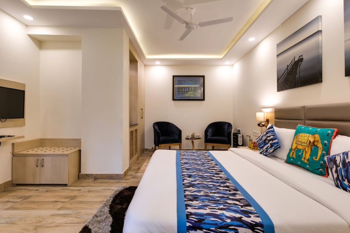 A hotel room with a bed, two chairs, a small table, a ceiling fan, and artwork on the walls.