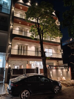 Un edificio de varios pisos con un coche estacionado adelante, un árbol en primer plano y múltiples balcones con iluminación.
