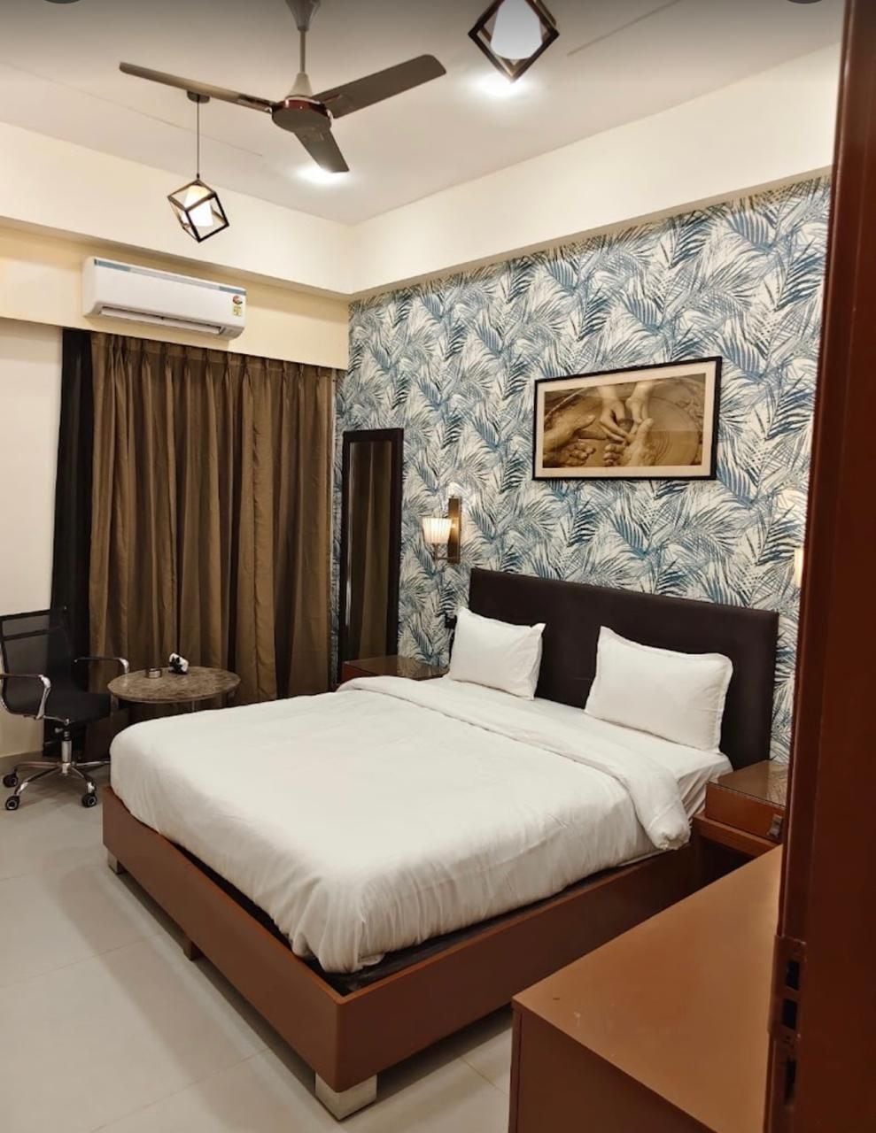 A hotel room with a bed, a desk, a chair, an air conditioning unit, a ceiling fan, and a wall with a tropical leaf pattern.