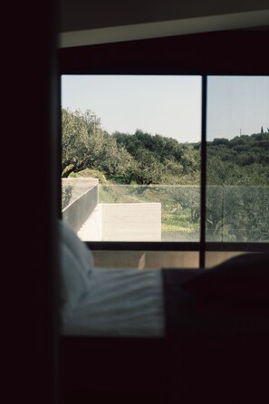 A room with a large window overlooking a landscaped area with trees and a concrete structure. - A Cube Villa in Keri (Zakynthos)