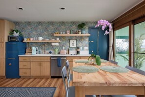 A modern kitchen with wooden cabinets, a built-in microwave, and a dining area with a wooden table and chairs. - Honoli‘i Sunrise House | 1BR/1BA | A More Better Home (Hilo)