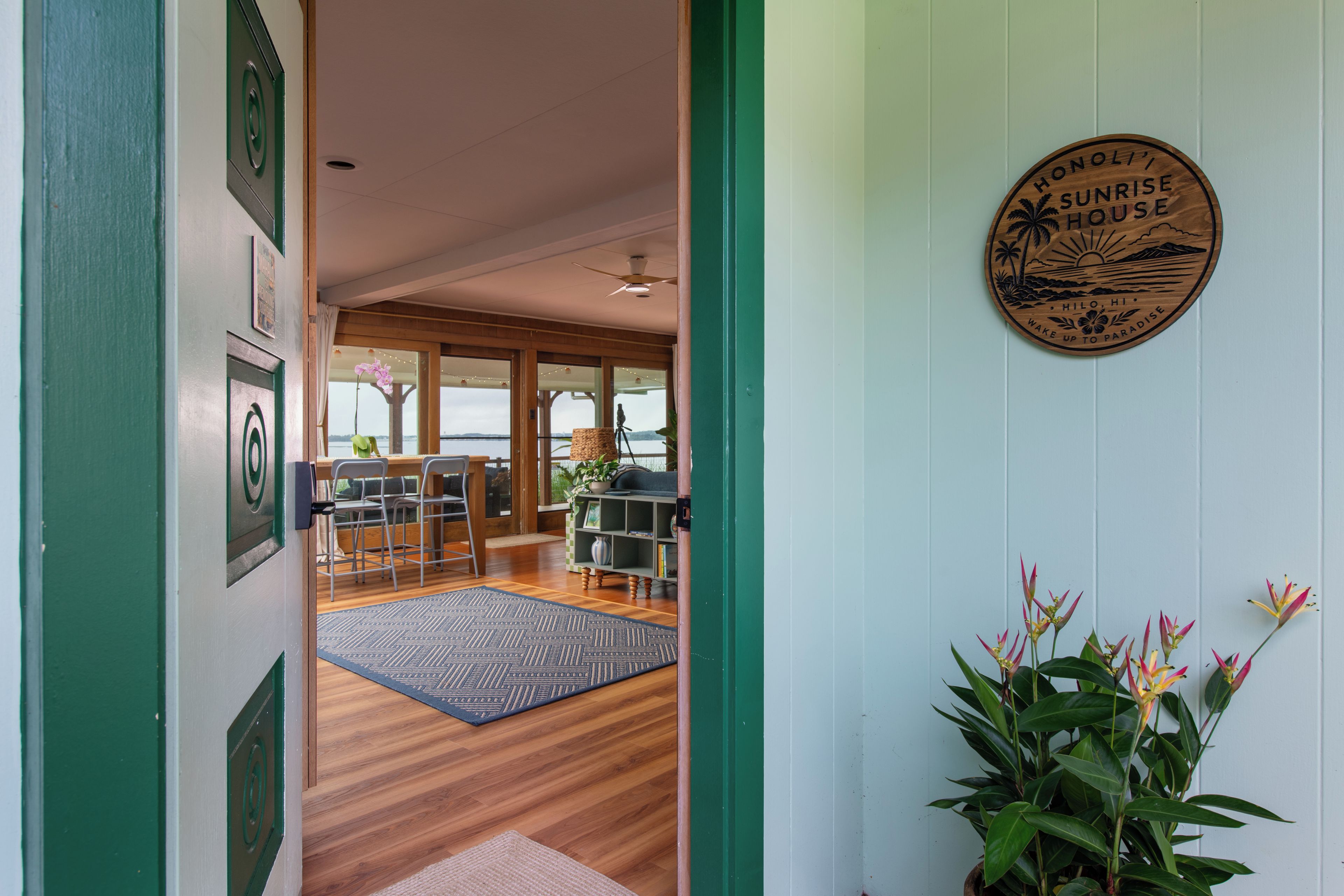 Un passage surmonté d’un cadre en bois mène à une pièce offrant une vue sur la mer, dotée d’un plancher en bois et d’un ventilateur de plafond.