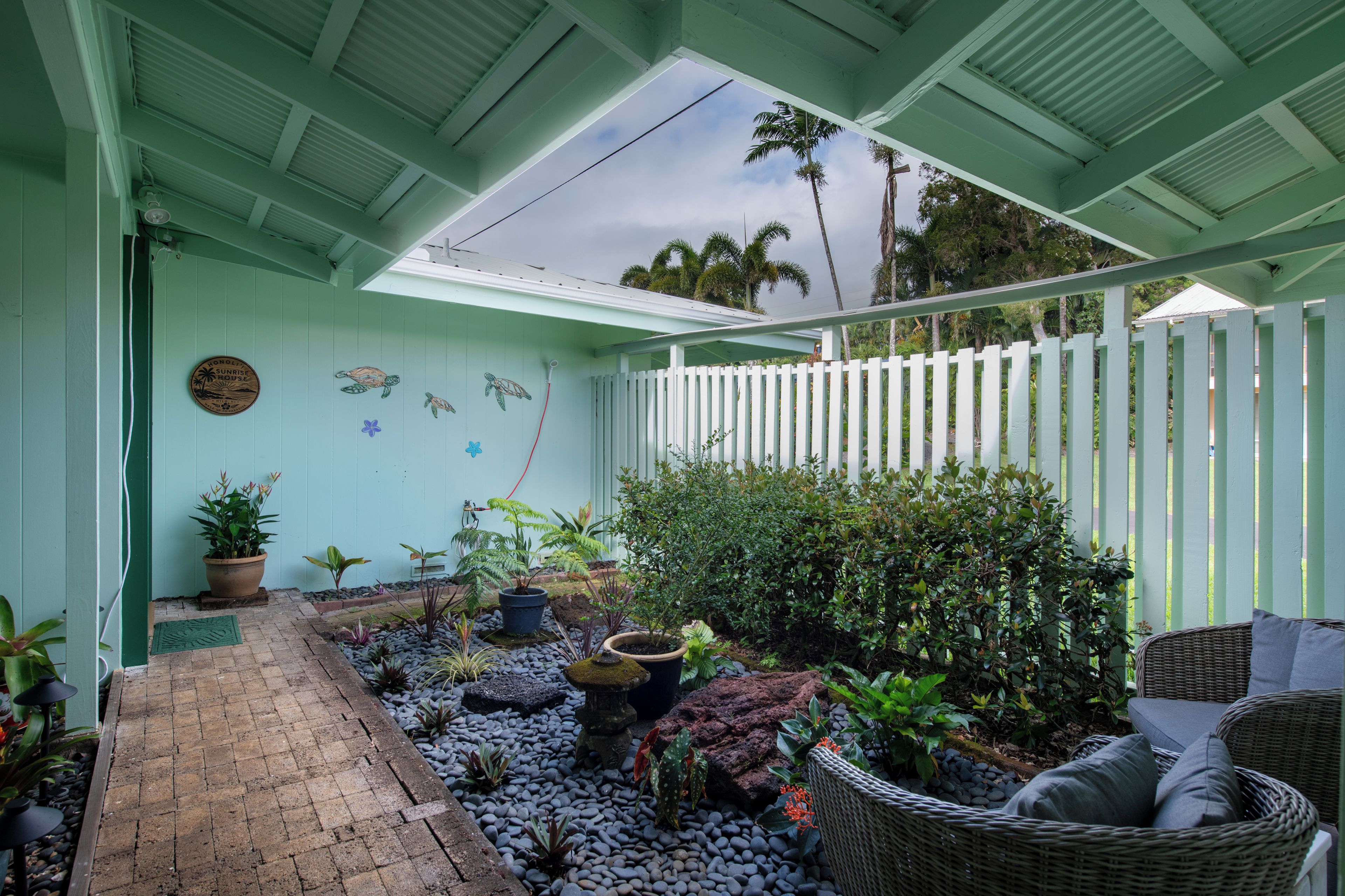 Un espace extérieur abrité, avec un chemin menant à un coin salon, entouré de plantes et d’une clôture blanche.