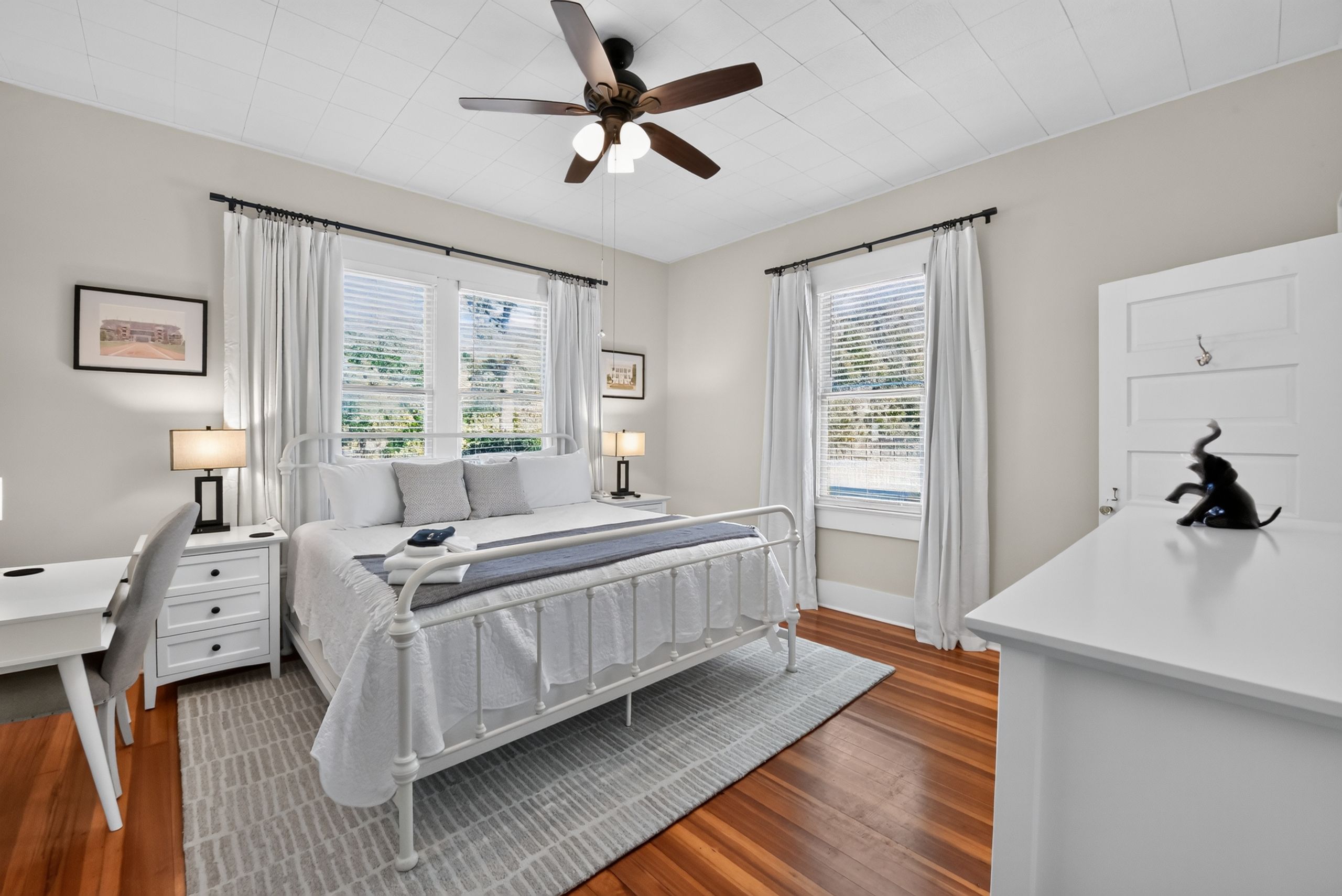 A bedroom with a bed, bedside tables, a desk, a ceiling fan, and windows with curtains.