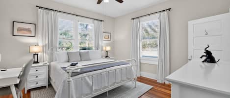 A bedroom with a bed, bedside tables, a desk, a ceiling fan, and windows with curtains.