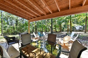 An outdoor covered patio with a glass table surrounded by chairs, overlooking a forested area.
