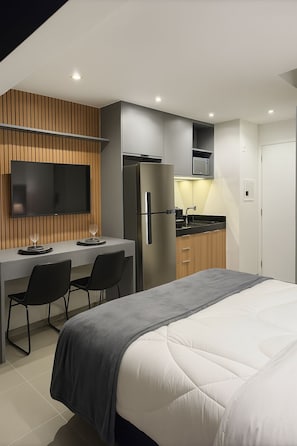 A modern hotel room with a bed, a desk with two chairs, a flat-screen TV mounted on the wall, a refrigerator, and a kitchenette. - Complete Studio Apartment in Jardins | Oscar Freire (São Paulo)