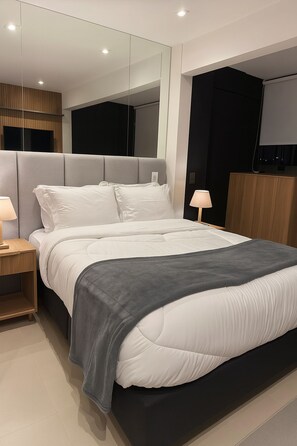 A modern bedroom with a large bed, bedside lamps, a mirror, and a television. - Complete Studio Apartment in Jardins | Oscar Freire (São Paulo)