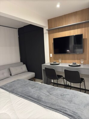 A modern hotel room with a bed, a desk, two chairs, a television, and a wooden panel wall. - Complete Studio Apartment in Jardins | Oscar Freire (São Paulo)