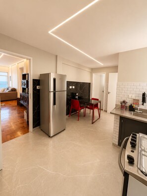 A modern kitchen with a stainless steel refrigerator, a dining area with red chairs, and a view of the living room. - Oceanfront - Panoramic View, Cinematic Sunset (Santos)