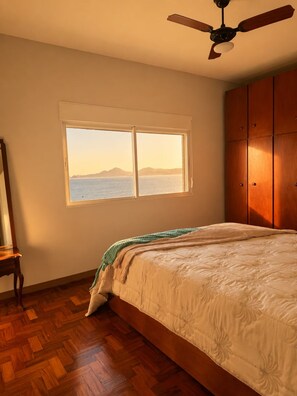 A bedroom with a bed, a window with a view of the sea, a ceiling fan, and wooden flooring. - Oceanfront - Panoramic View, Cinematic Sunset (Santos)