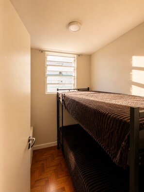 Quarto pequeno com uma cama de solteiro, beliche, janela com cortinas e piso de madeira.
