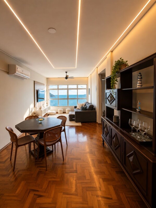 Uma sala de estar moderna com piso de madeira em padrão espinha de peixe, uma área de jantar com mesa de madeira escura e cadeiras de vime, uma janela grande com vista para o mar, e uma estante embutida com objetos decorativos.