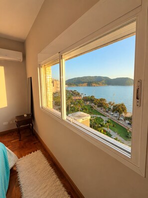 A room with a large window overlooking a coastal area with a view of the sea and hills. - Oceanfront - Panoramic View, Cinematic Sunset (Santos)