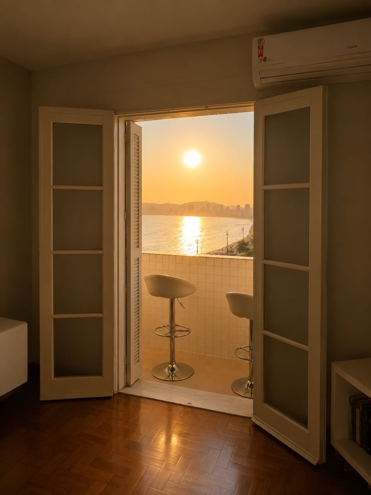 Une porte ouverte donnant sur un balcon avec vue sur un coucher de soleil au-dessus d’une étendue d’eau, deux tabourets de bar et un appareil de climatisation.