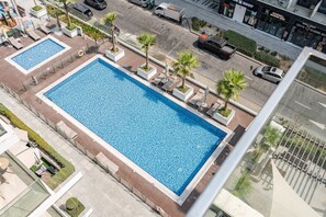 Ein großer rechteckiger Pool mit Kachelmuster, umgeben von Liegestühlen und Palmen, in einer städtischen Umgebung mit Gebäuden und einer Straße im Hintergrund.