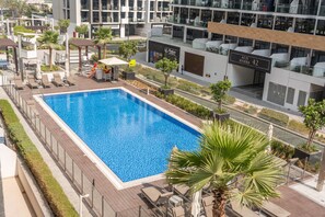 Ein rechteckiger Swimmingpool mit glasklar blauem Wasser, umgeben von Liegestühlen und Sonnenschirmen, mit Blick auf moderne Gebäude und eine Straße im Hintergrund.