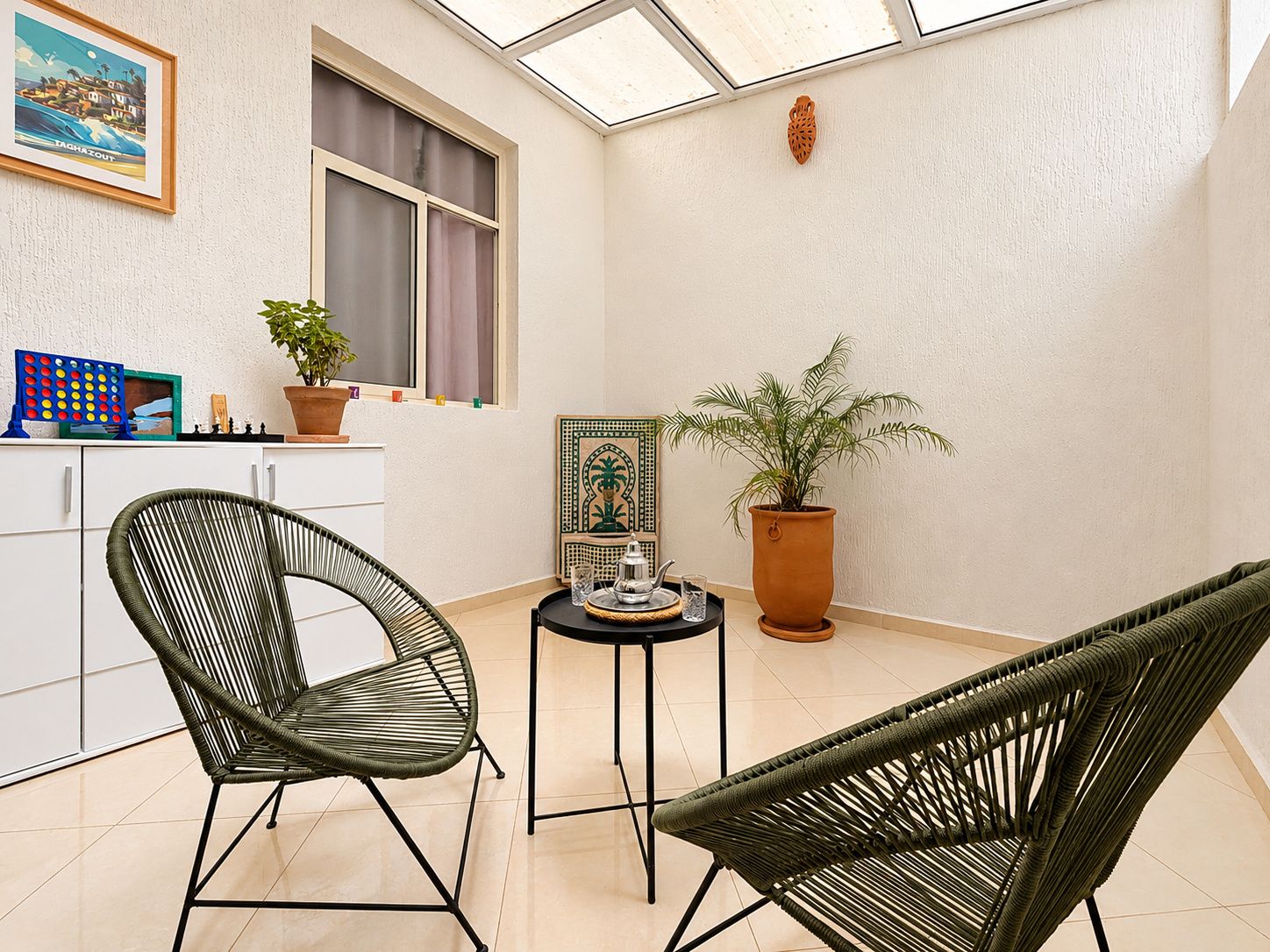 Een kamer met twee groene rieten stoelen, een kleine tafel met een plant, een raam en een aan de muur hangend schilderij.