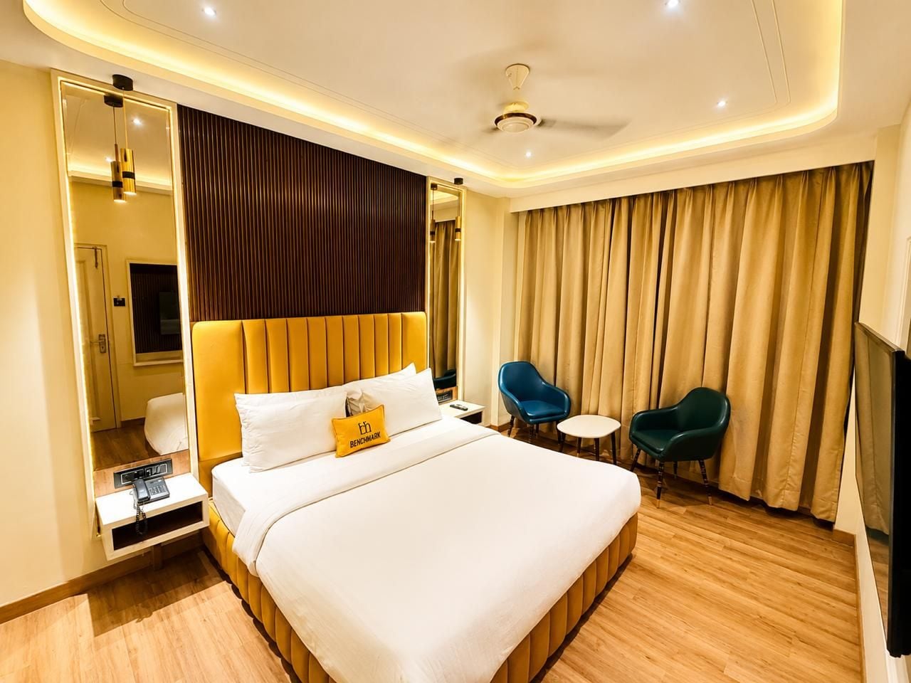 A hotel room with a large bed, bedside tables, a desk, two chairs, a ceiling fan, and a large window with curtains.