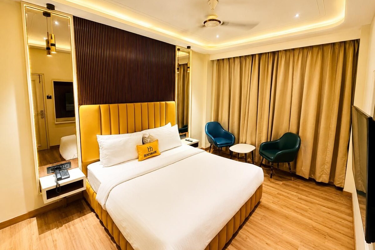 Quarto de hotel com uma cama grande, mesas de cabeceira, escrivaninha, duas cadeiras, um ventilador de teto e uma janela ampla com cortinas.