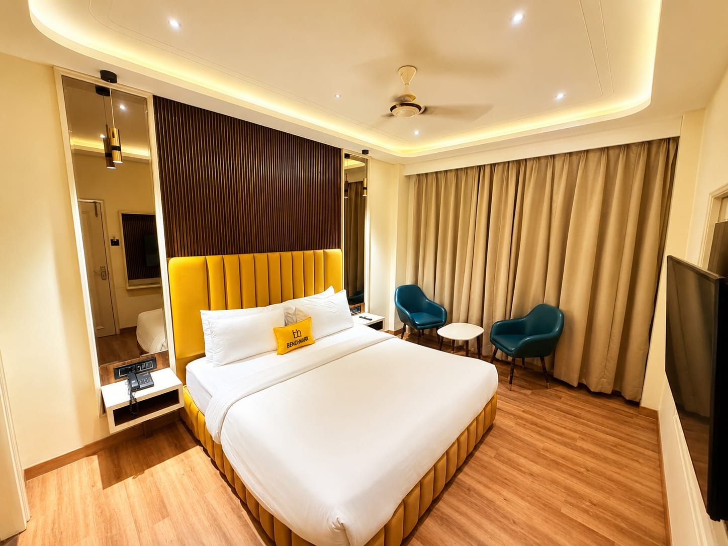 A hotel room with a large bed, a desk with a telephone, two chairs, a television, and a ceiling fan.