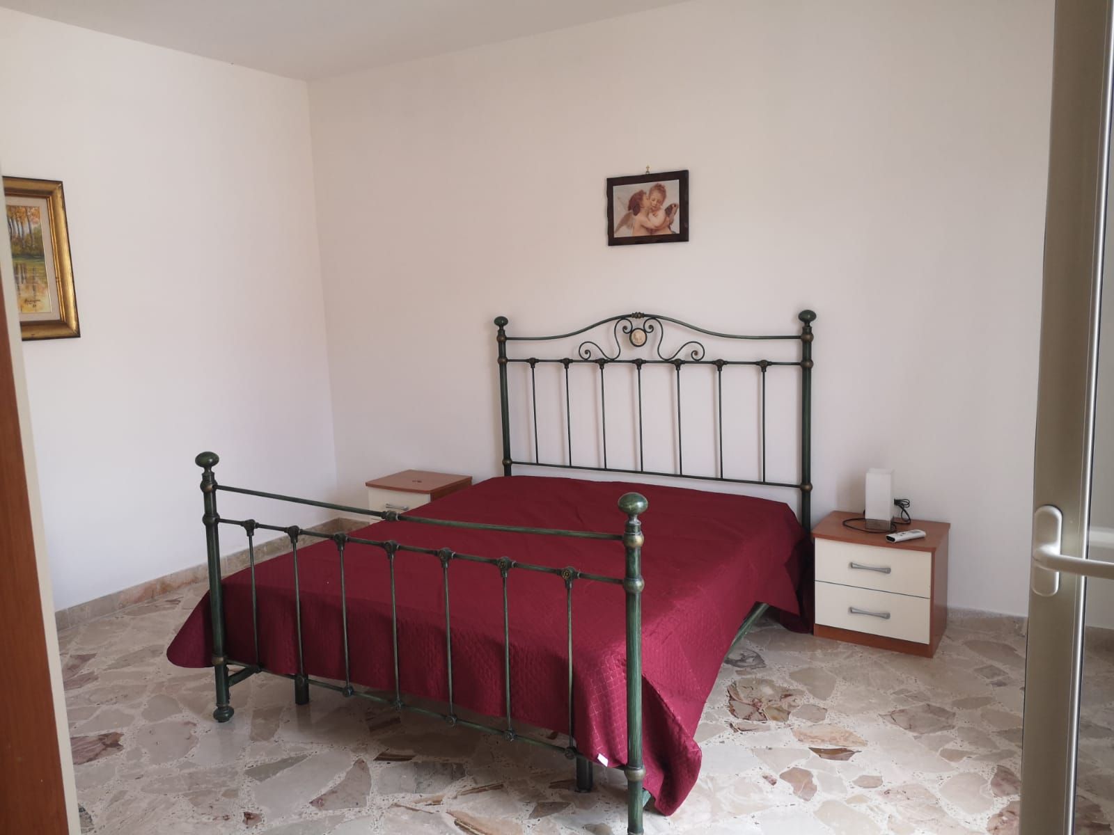 Una camera da letto con una struttura del letto in metallo, una coperta rossa, un comodino, un cassetto, un quadro appeso al muro e una porta.
