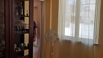 Una stanza con pavimento in pietra, un armadio in legno con ante in vetro, una ventola a piantana e una finestra con tende leggere.