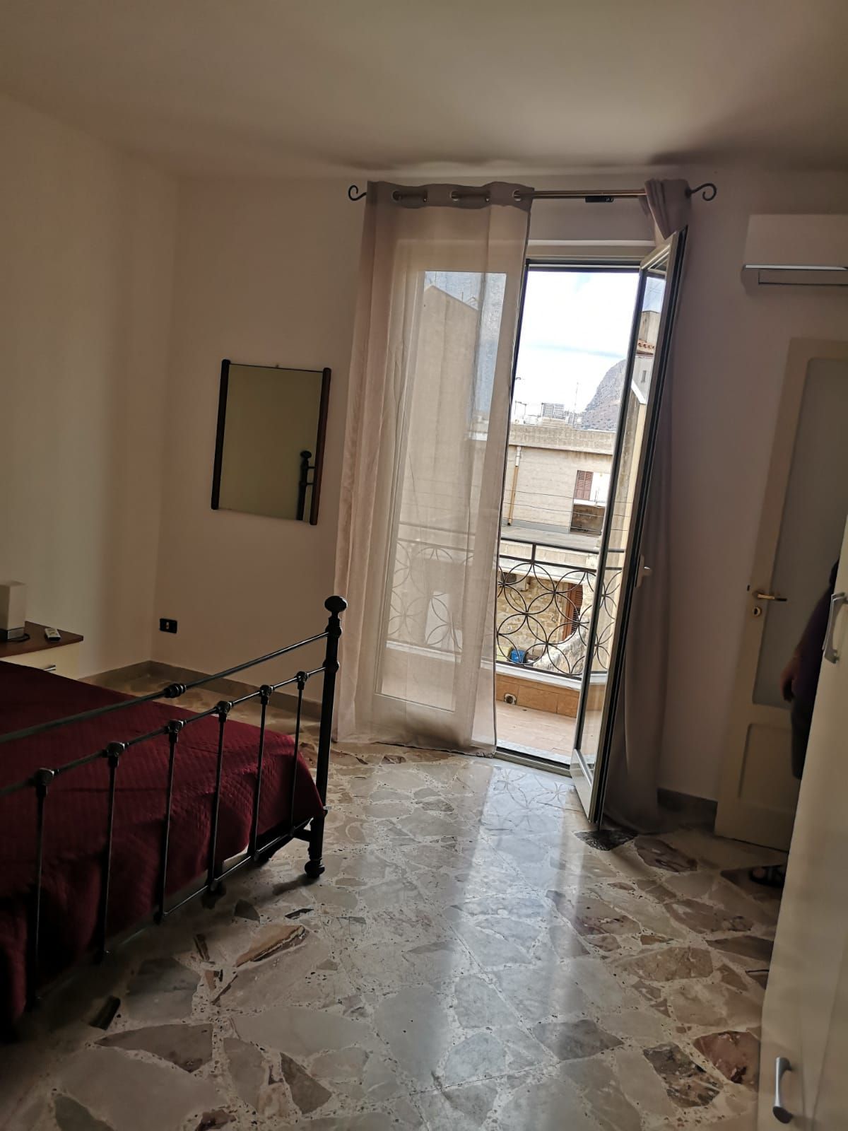 Ein Schlafzimmer mit einem Bett, einem Spiegel, einer Tür zu einem Balkon und Blick auf Gebäude.