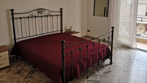 A bedroom with a metal bed frame, a red bedspread, a bedside table with a lamp, a framed picture on the wall, and a window with a view of the outside. - 5 minutes from the square and the airport (Terrasini)