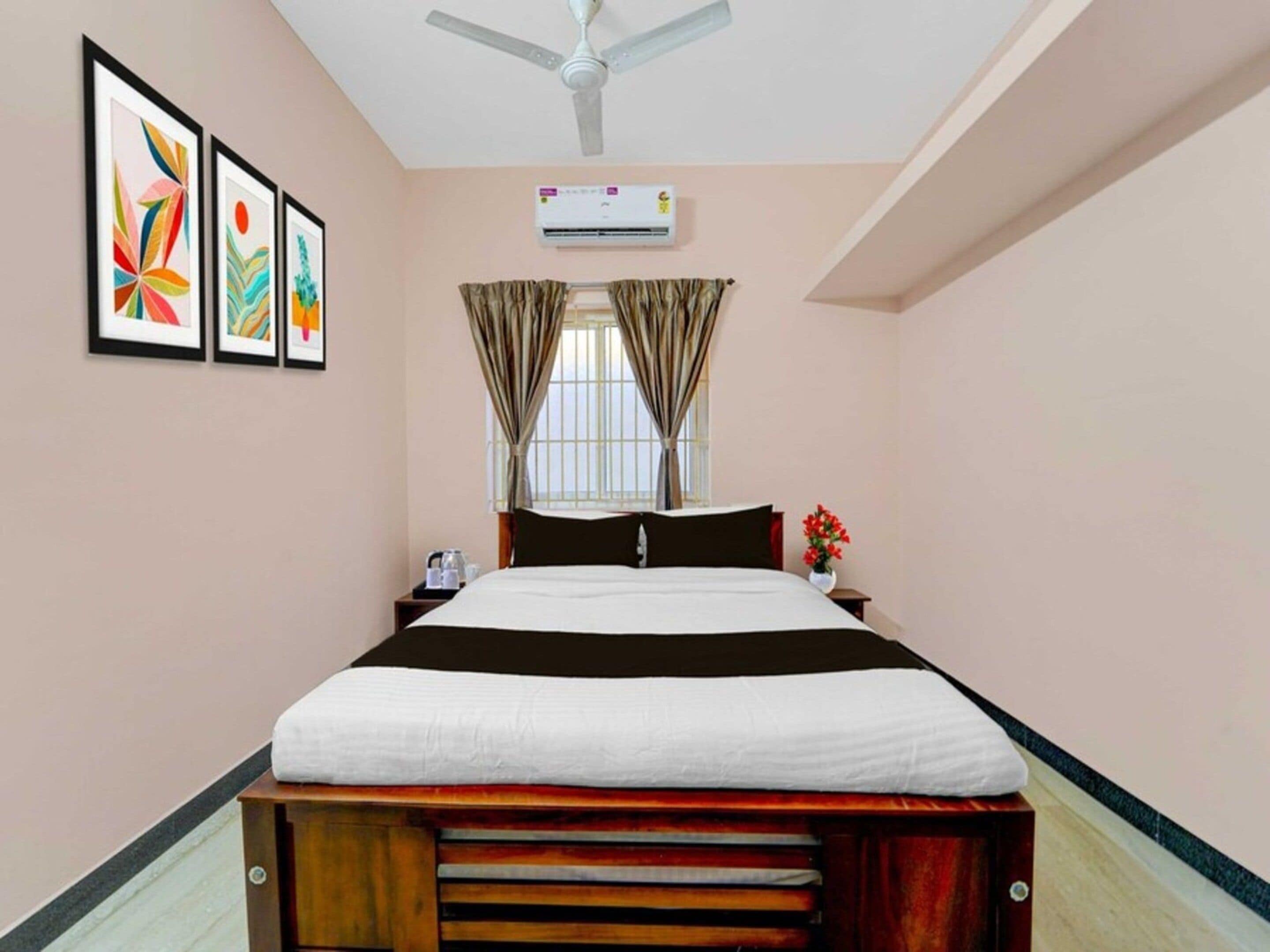 A hotel room with a bed, a ceiling fan, an air conditioning unit, a window with curtains, and framed artwork on the wall.