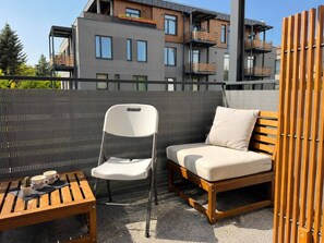 Ein Sitzbereich im Freien mit Stuhl und Sofa, einem kleinen Tisch mit Tasse und Pflanze sowie Blick auf ein modernes Gebäude mit Balkonen.