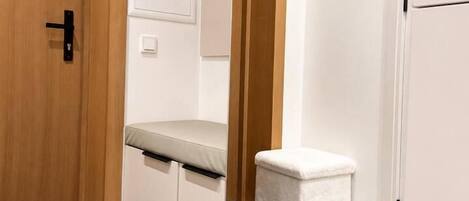 Un couloir avec une porte en bois, un banc blanc, un petit tabouret blanc et un tapis noir sur le sol.