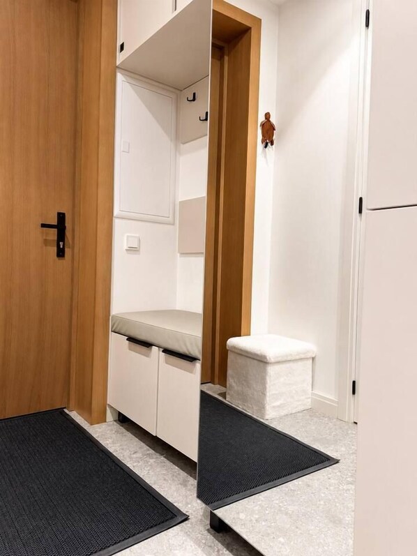 A hallway with a wooden door, a white bench, a small white stool, and a black mat on the floor.