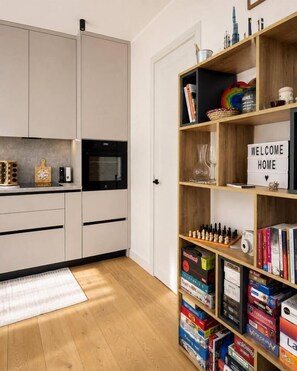 Eine moderne Küche mit einem eingebauten Ofen, einem Holzregal voller Bücher und Dekoration, und einem Schild mit der Aufschrift 'WELCOME HOME' an der Tür.