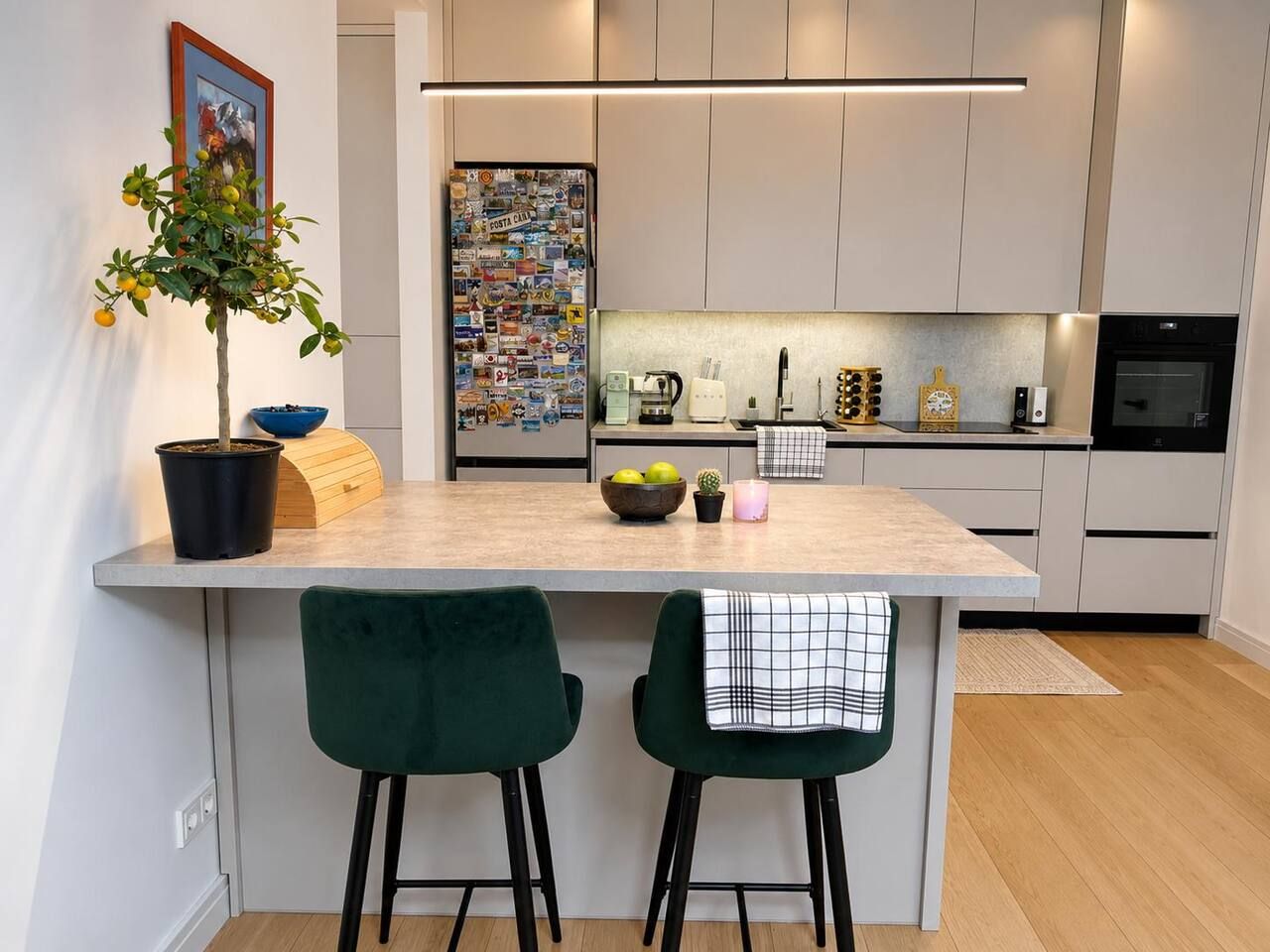 A modern kitchen with a central island, two green bar stools, a potted plant, and a refrigerator covered with magnets.
