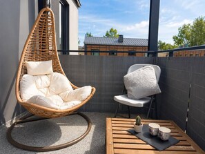 A balcony with a wicker chair, a white chair, and a small table with a cactus and two cups.