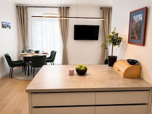 A modern kitchen with a central island, dining area with chairs, a flat-screen TV, and a potted plant.