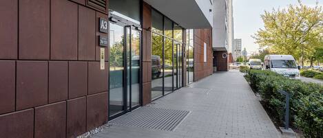 Um edifício moderno com entrada de vidro, calçada pavimentada e área de estacionamento com veículos.