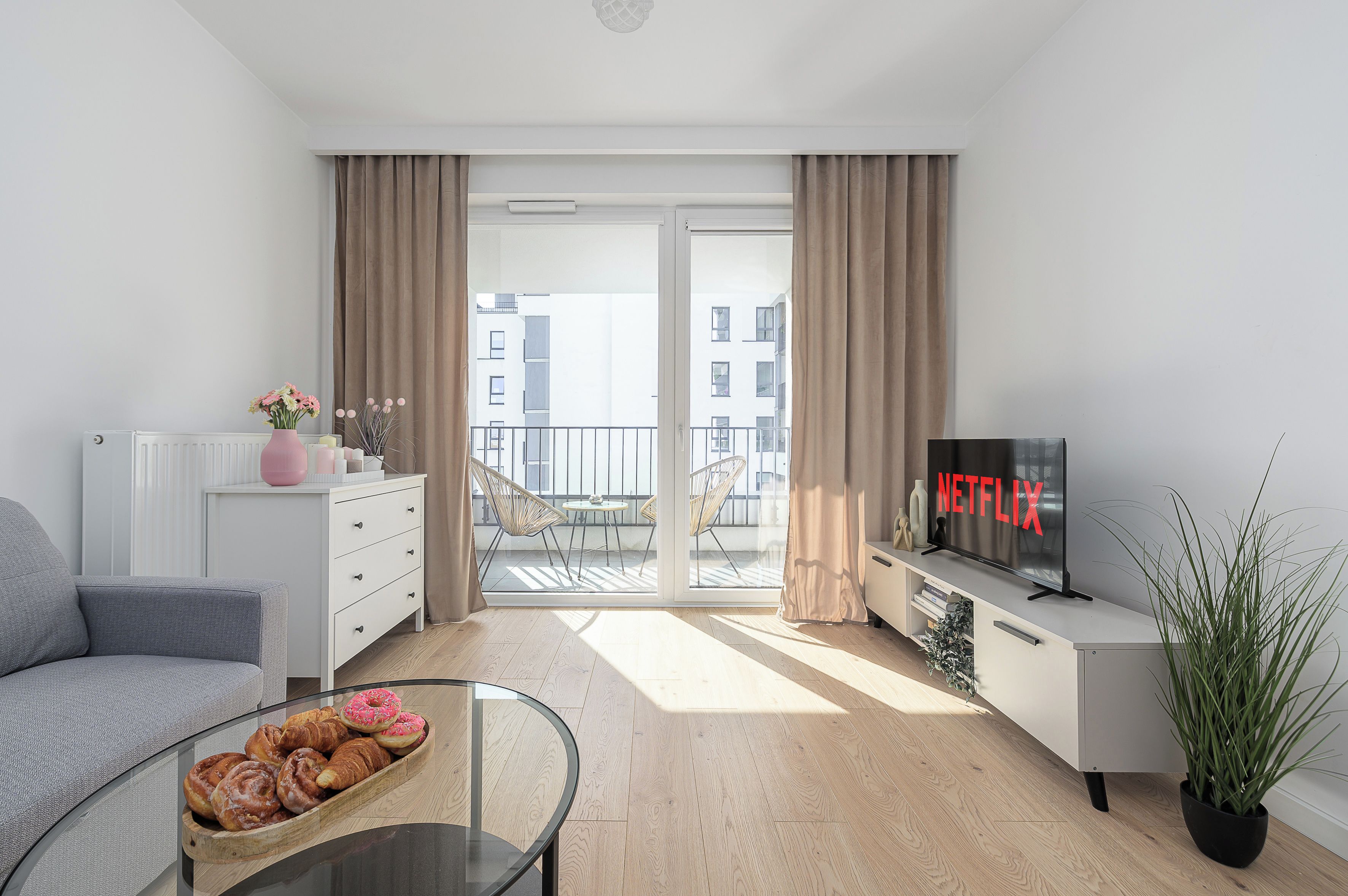 A modern living room with a sofa, a coffee table with pastries, a television, and a balcony with a view of buildings.