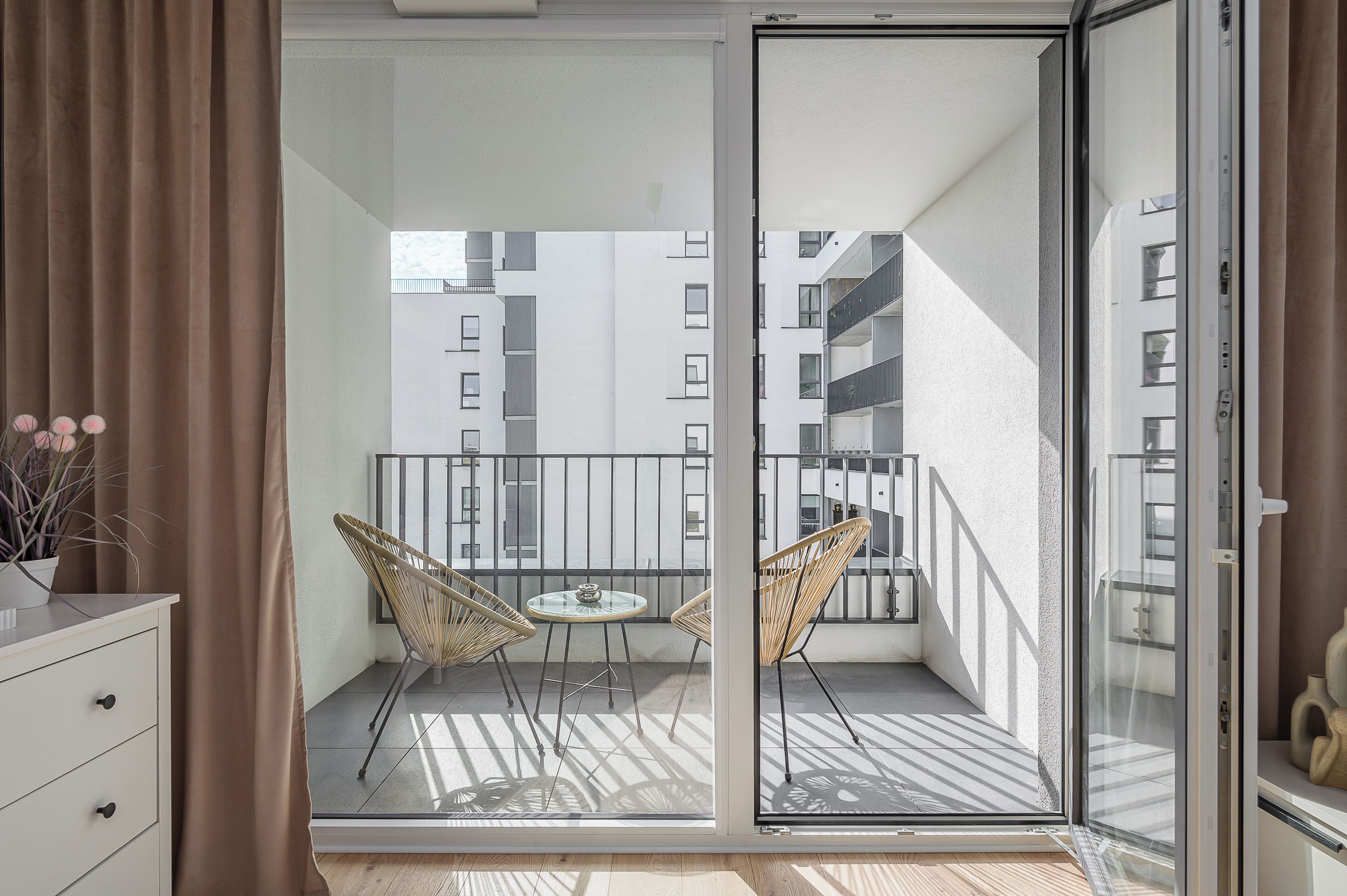 A balcony with two wicker chairs and a small table, overlooking a cityscape with multiple buildings.