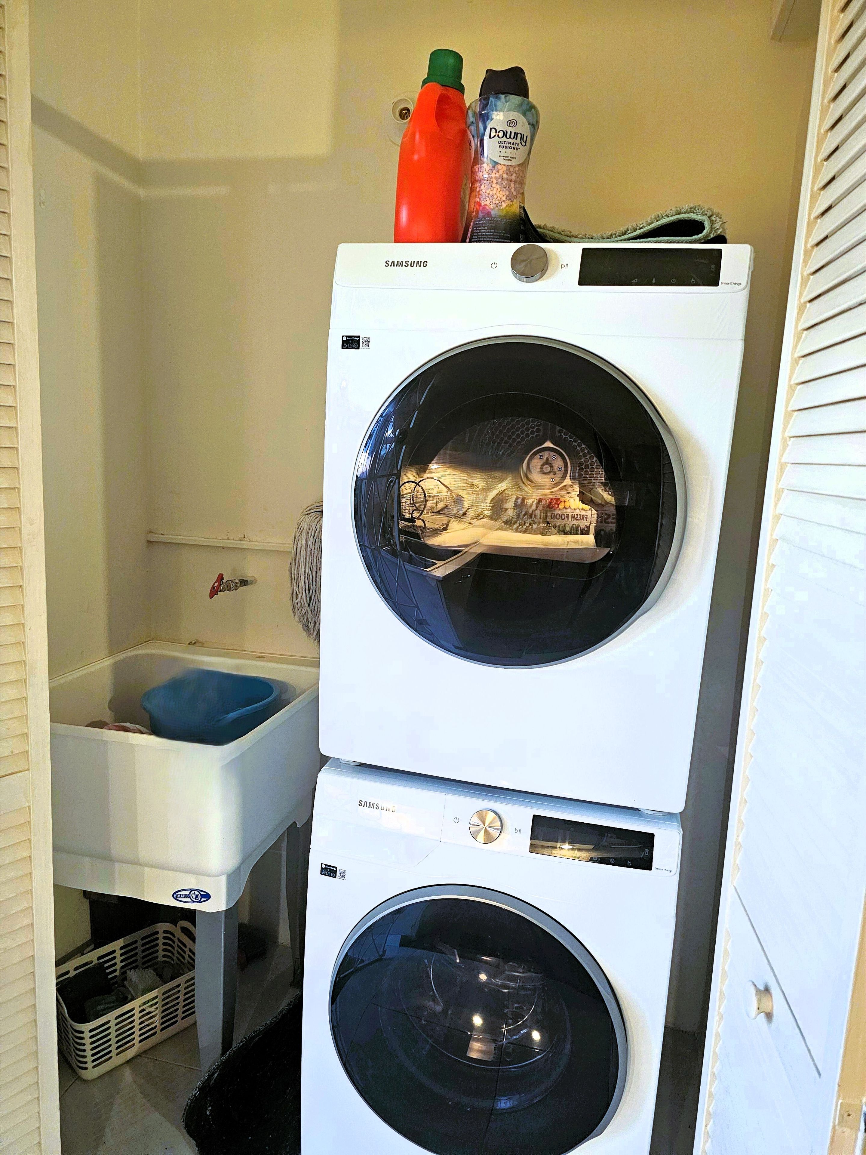 Un área de lavandería con dos lavadoras blancas apiladas, un lavabo azul y un estante blanco con productos de limpieza.