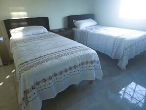 A hotel room with two beds, each with a patterned bedspread and pillows, a nightstand with a remote control, and a window providing natural light. - Camelot Blue - A Tranquil Oceanview Escape (Discovery Bay)