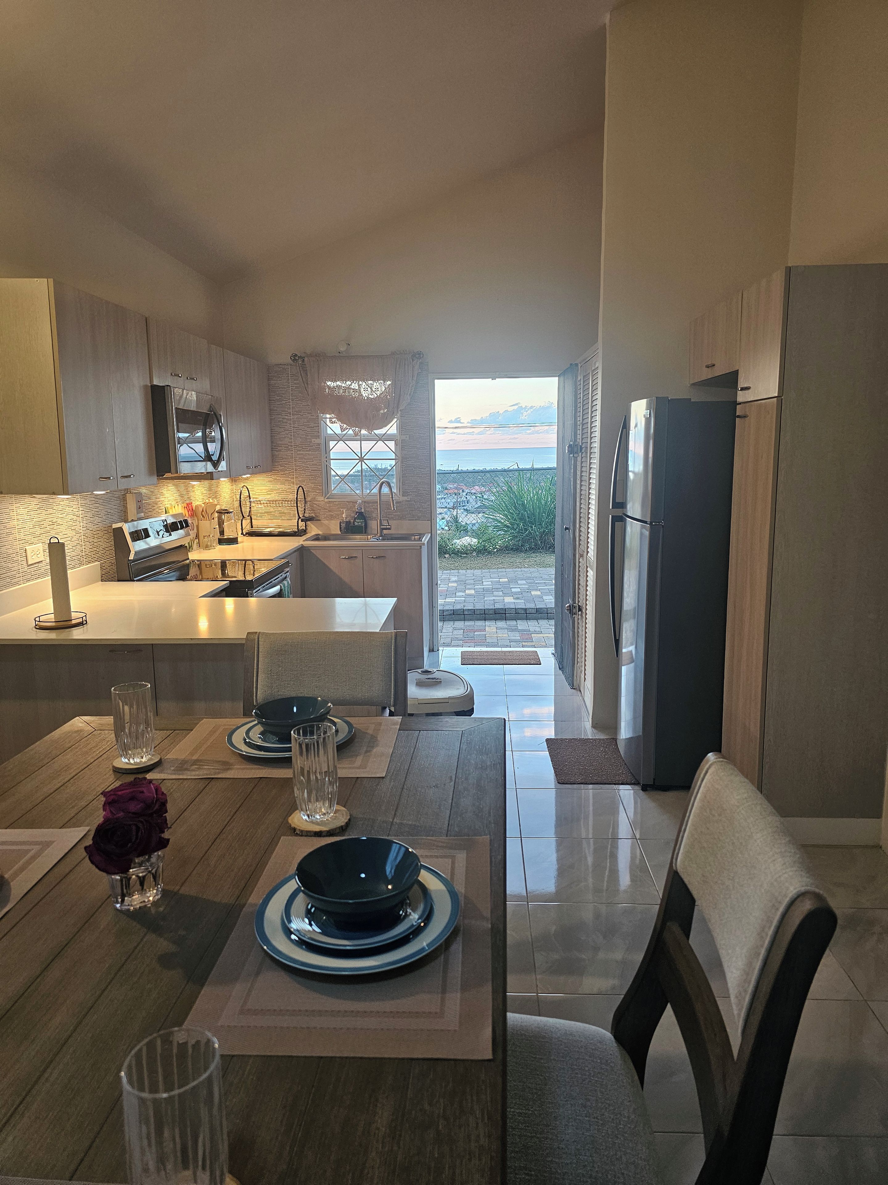 A modern kitchen with a view of the sea, a dining area with a table set for two, and a refrigerator.