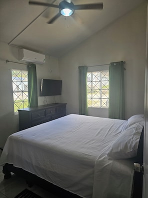 Ein Schlafzimmer mit Bett, Fernseher, Deckenventilator und einem Fenster mit Vorhängen.