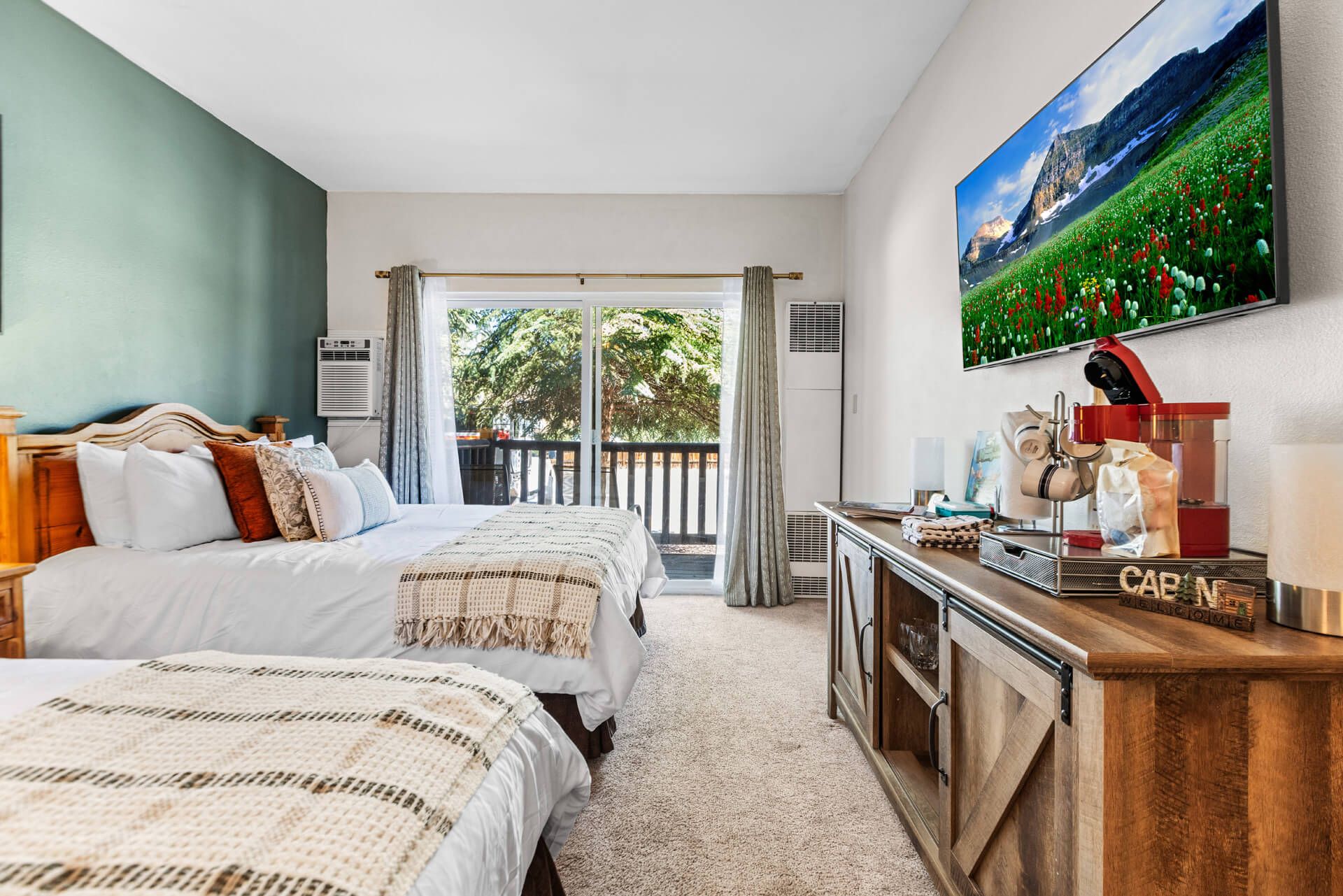 Habitación de hotel con dos camas, un escritorio de madera, un cuadro y vista a un jardín a través de un ventanal.