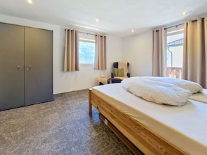 Ein Schlafzimmer mit Bett, Kleiderschrank, Fenster mit Vorhängen und Blick ins Freie.